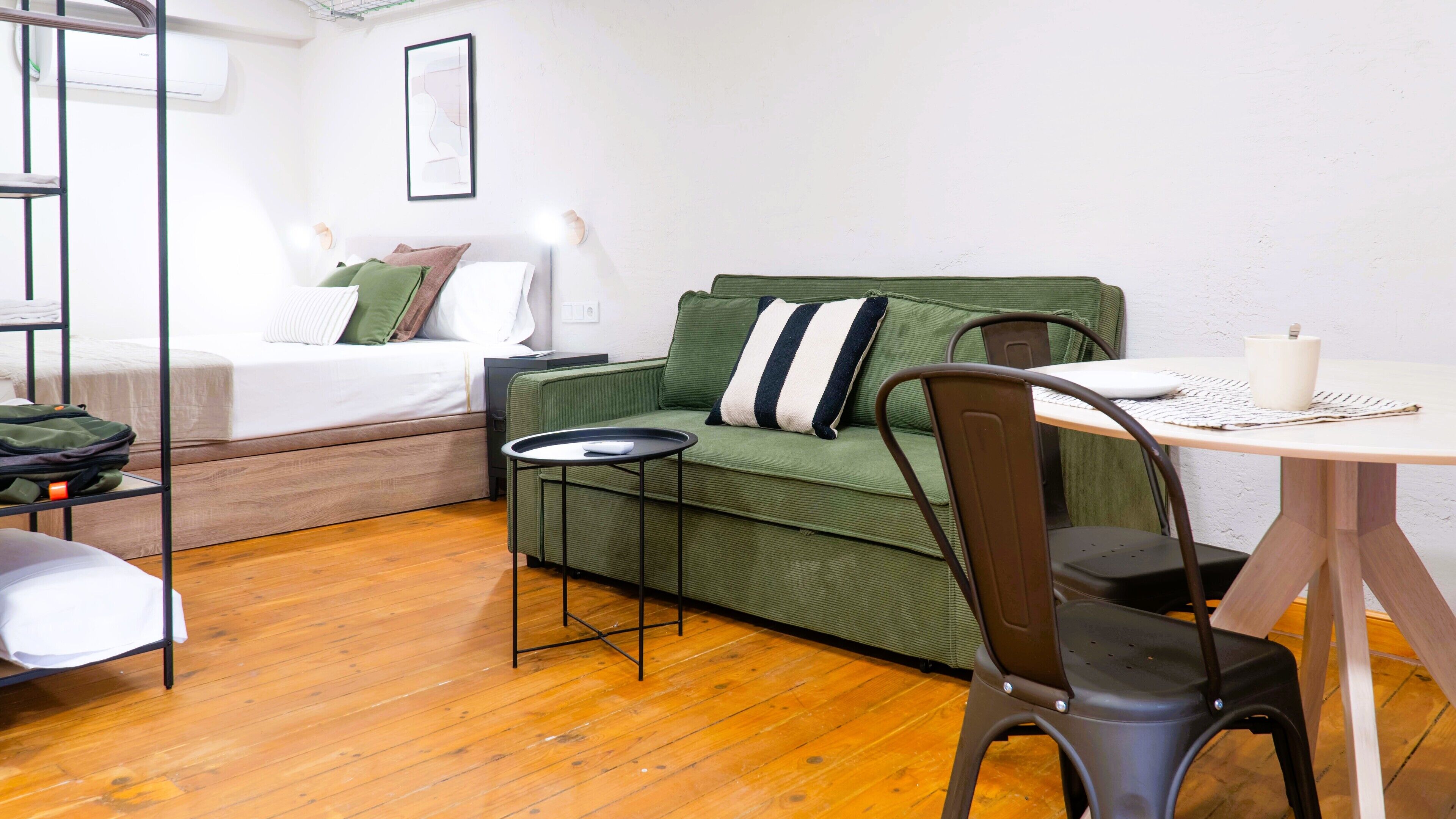A compact room with a green sofa, a small round table, a chair, a bed with a white mattress, and a wall-mounted air conditioner.