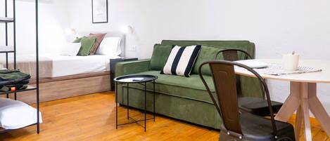 A compact room with a green sofa, a small round table, a chair, a bed with a white mattress, and a wall-mounted air conditioner.
