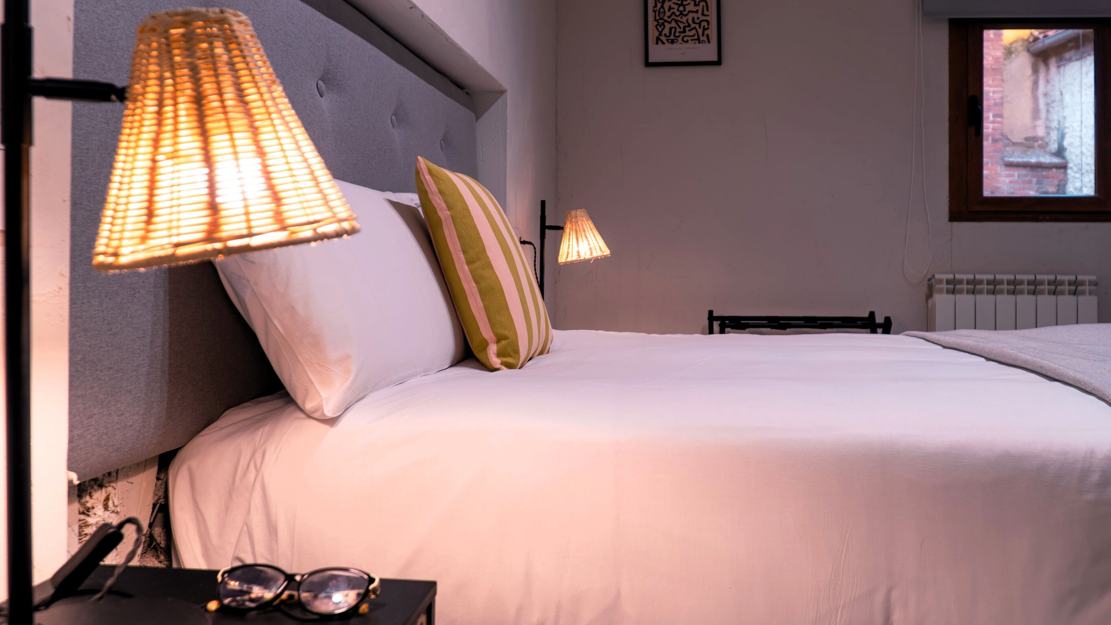 Una cama bien hecha con ropa blanca, dos almohadas y una mesita de noche con vasos y una lámpara.