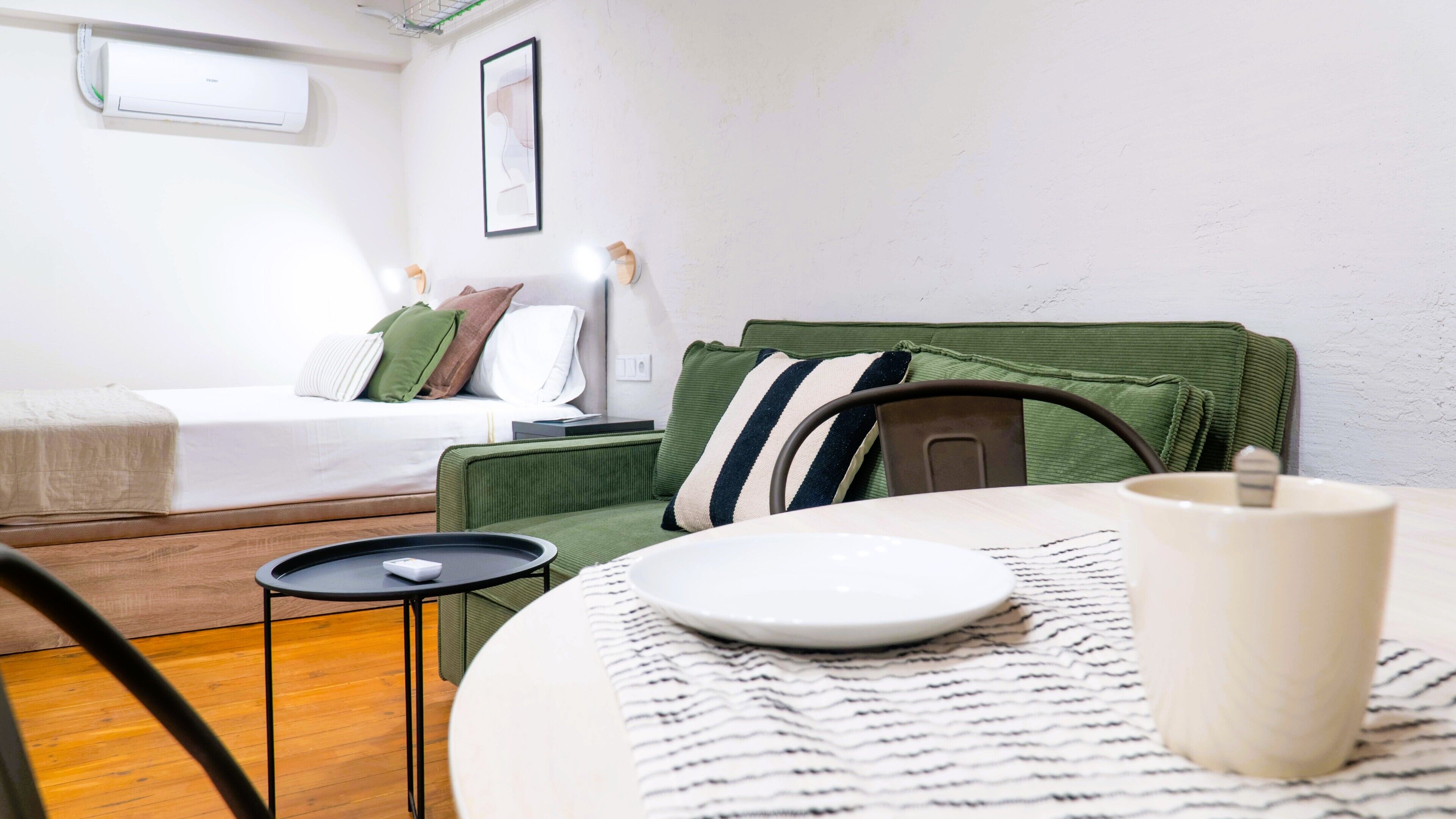 A hotel room with a bed, a green sofa, a small table with a plate and a cup, and a wall-mounted air conditioner.
