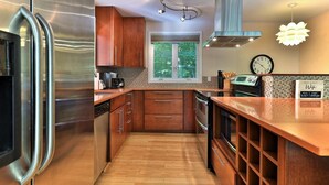 A modern kitchen with stainless steel appliances, wooden cabinets, a window with a view, and a clock on the wall.