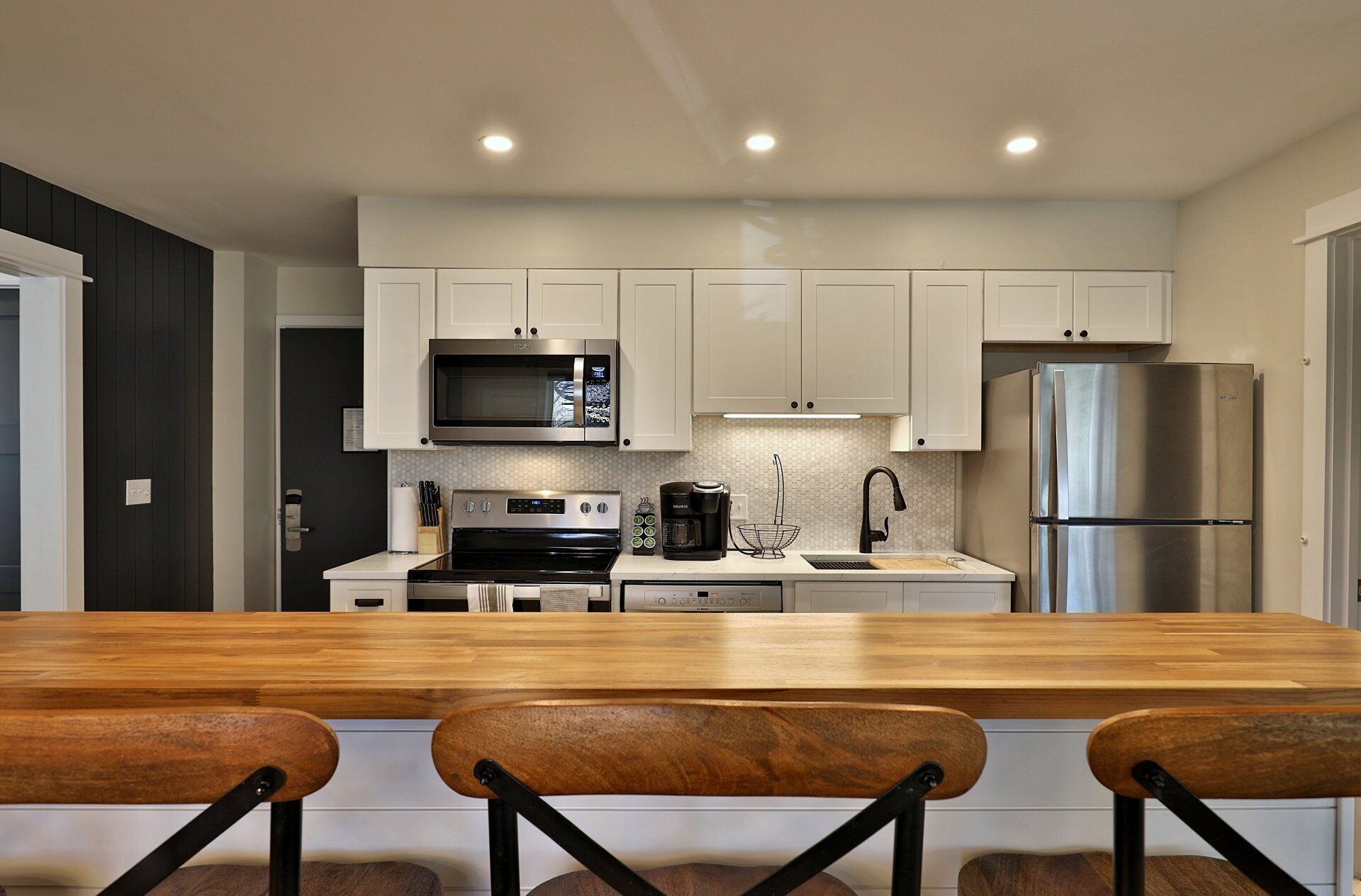 A modern kitchen with white cabinets, a stainless steel refrigerator, a microwave, and a coffee maker. There is a wooden dining table with two chairs in the foreground.