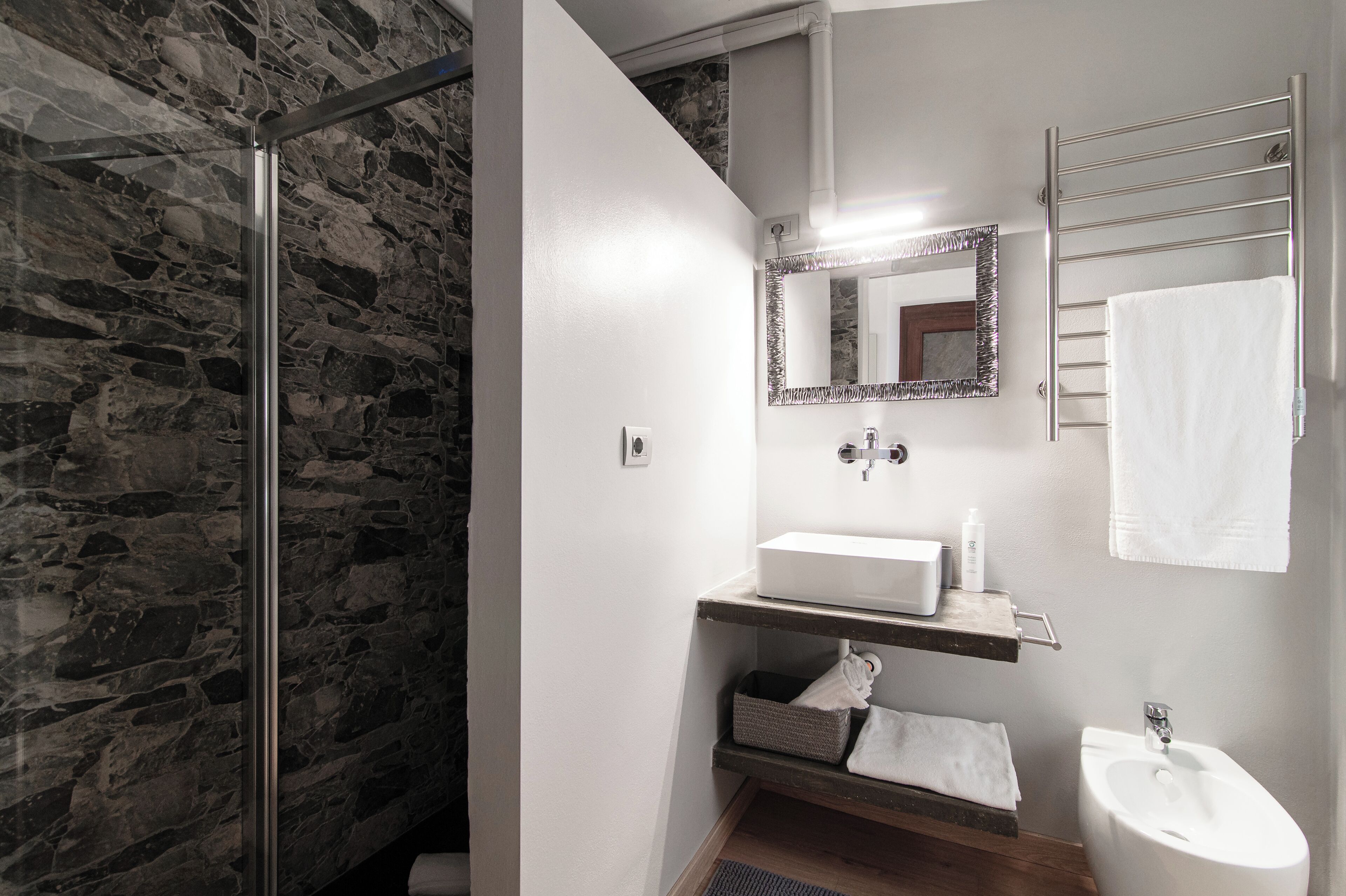 A bathroom with a stone wall, a glass shower enclosure, a white sink, a mirror, and a towel rack.