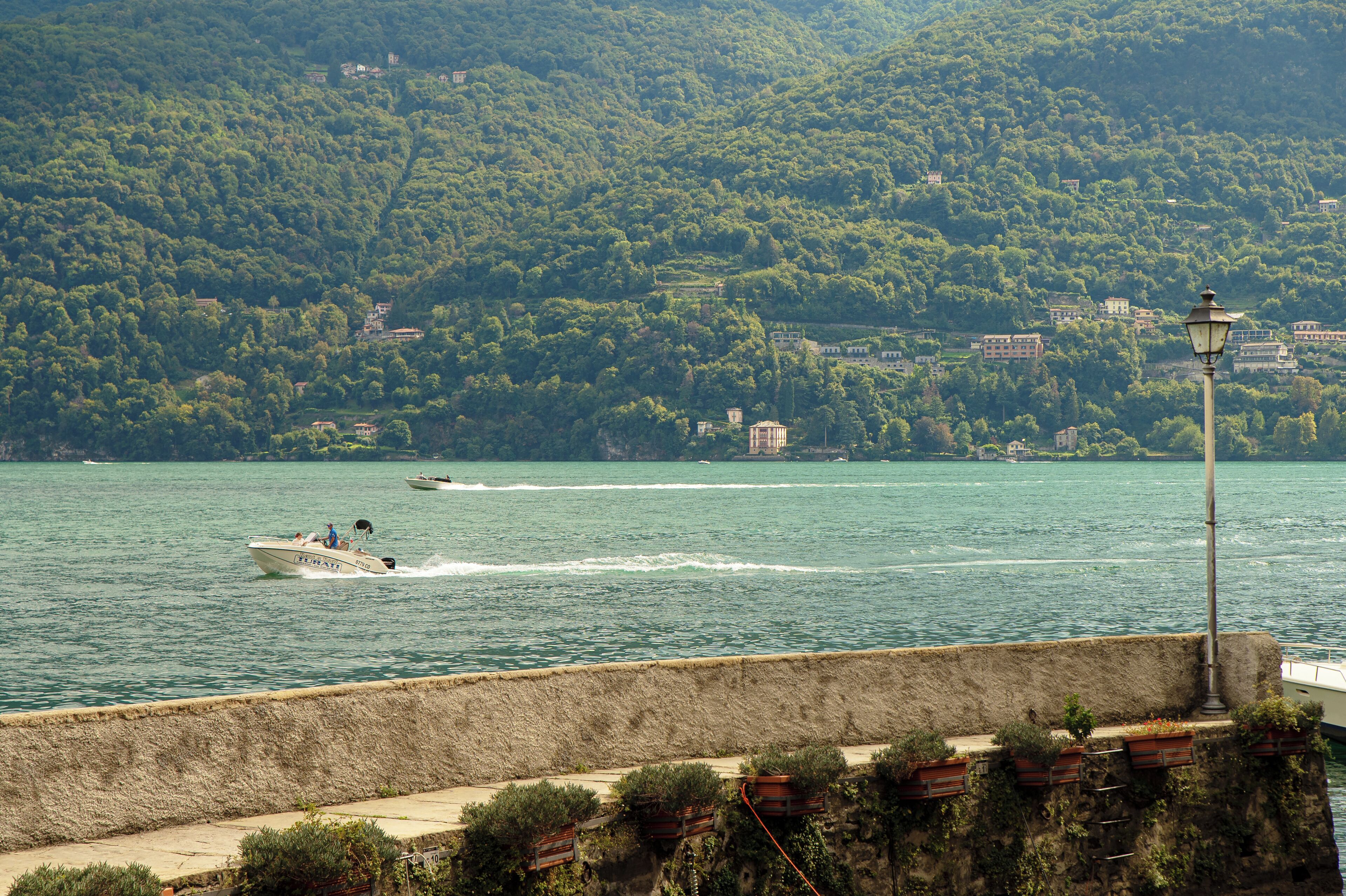 A boat speeding across a lake with a backdrop of a forested hillside and a stone wall with potted plants.