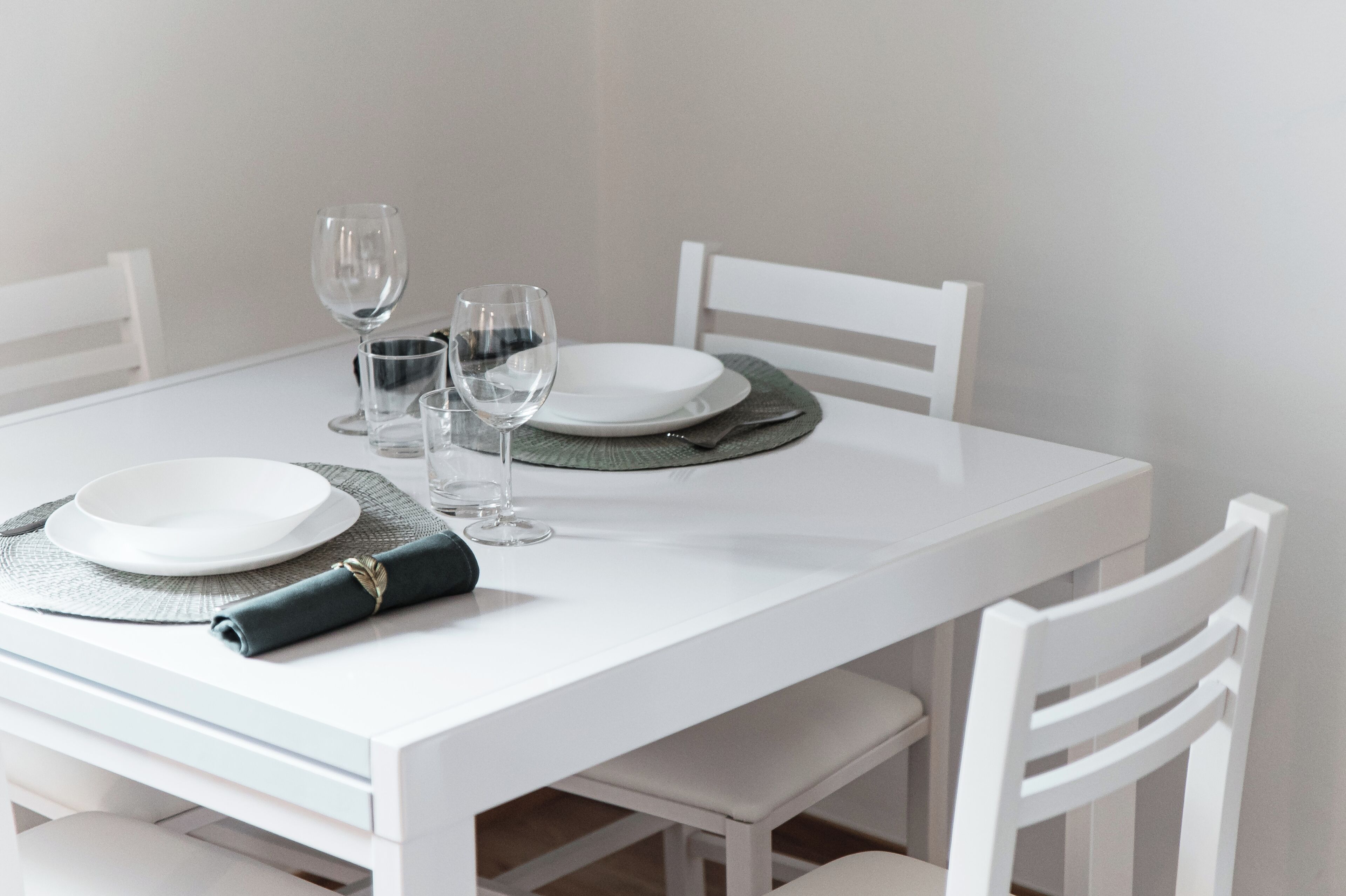 A white dining table with four white chairs, a folded napkin, and two wine glasses.