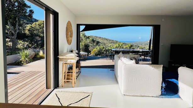 Un salon moderne avec un canapé blanc, une table en bois et une vue sur une terrasse et un paysage naturel.