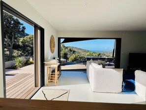 A modern living room with a white sofa, a wooden table, and a view of a deck and natural landscape. - Architect-designed villa with sea and scrubland views (Figari)