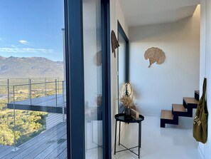 Panoramic mountain views from a spacious room with a minimalist design, featuring a black table, vase with dried plants, and woven wall hanging.