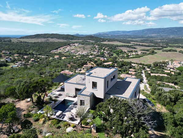 A modern house with a swimming pool, surrounded by lush greenery and hills in the background.