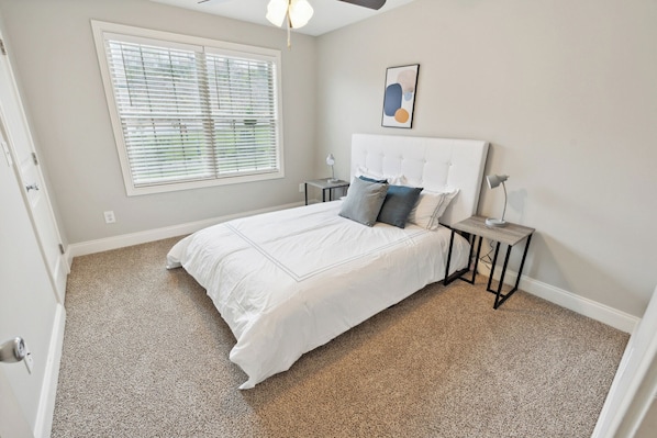 A bedroom with a large bed, a window with blinds, a bedside table with a lamp, and a framed picture on the wall.