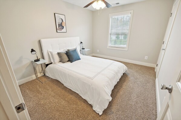A bedroom with a white bed, a nightstand, a lamp, a window with blinds, and a ceiling fan.