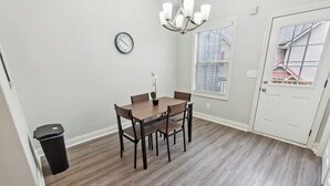 A dining area with a wooden table, chairs, a potted plant, a wall clock, and a window with blinds.