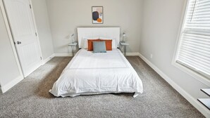 Un dormitorio con una cama blanca, dos mesitas de noche, una puerta blanca y un ventanal con persianas.