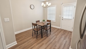 A dining area with a wooden table and chairs, a chandelier, a clock, and a door with blinds.