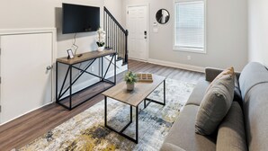 A modern living room with a sofa, a coffee table, a TV mounted on the wall, a staircase, and a door.