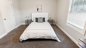 A bedroom with a large bed, two bedside tables, a nightstand, a window with blinds, and a door.
