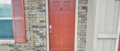 A red door with the number 6, a brick wall, and a window with shutters.