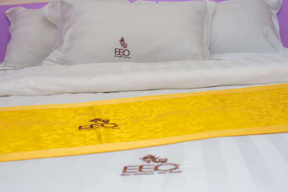 A bed with white bedding and a yellow bedspread with the EEO logo.