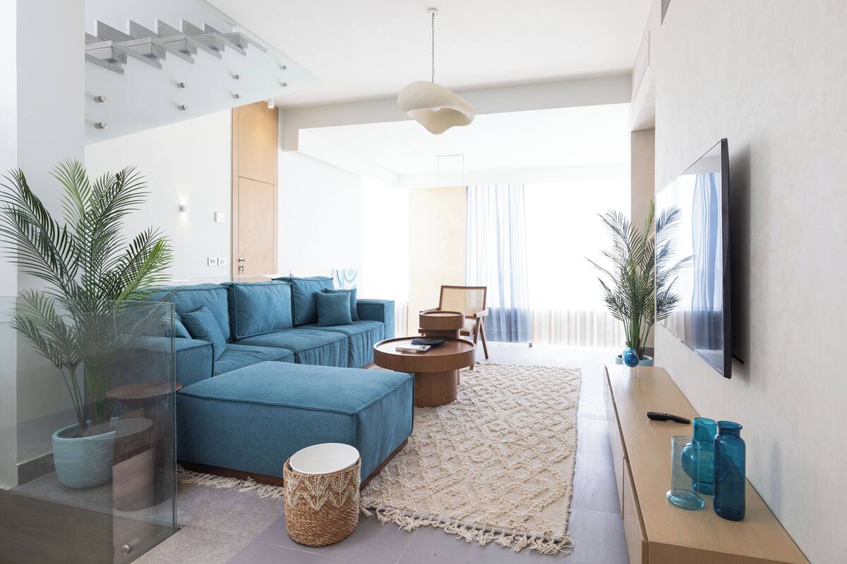 Een moderne woonkamer met een blauwe hoekbank, een ronde salontafel en een flatscreen televisie aan de muur.