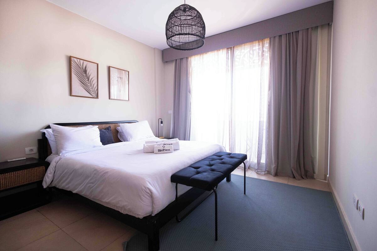Een opgemaakt bed met witte lakens, een nachtkastje, een bankje en een hanglamp.