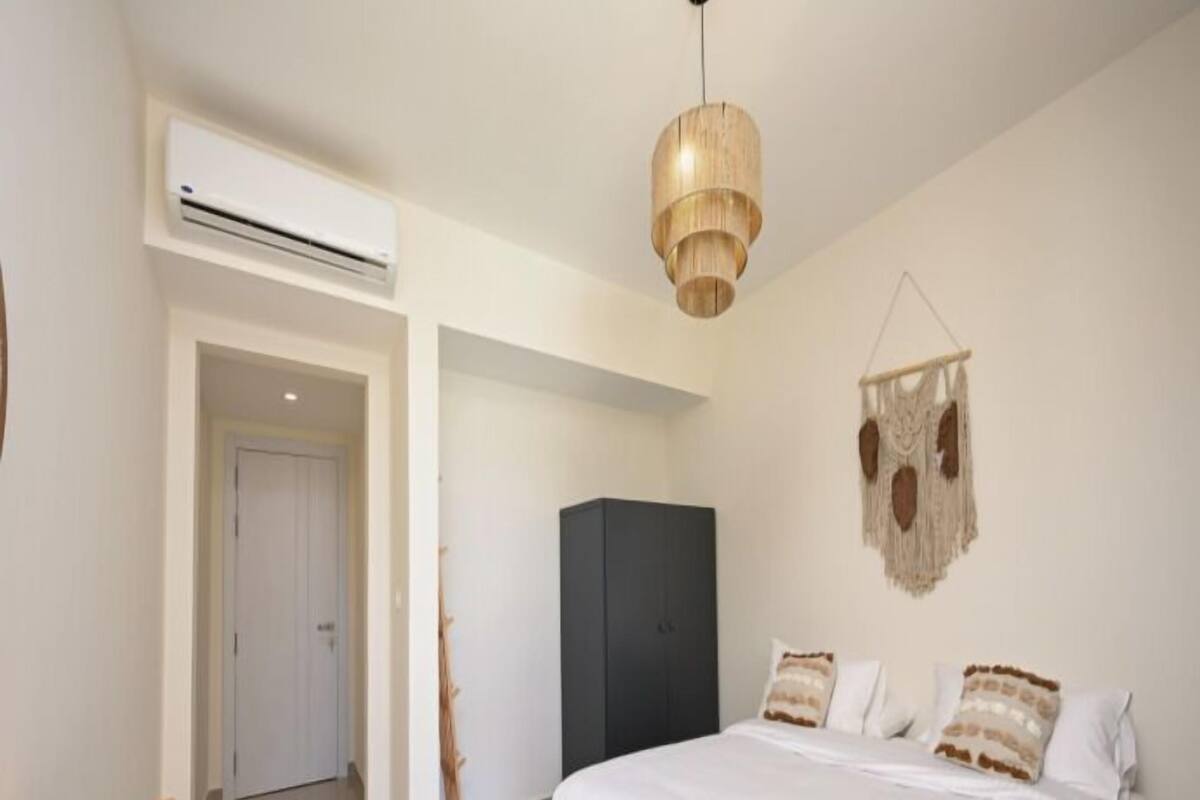Een slaapkamer met een bed, een houten tafel, een airconditioner, een kledingkast en een macramé wanddecoratie.
