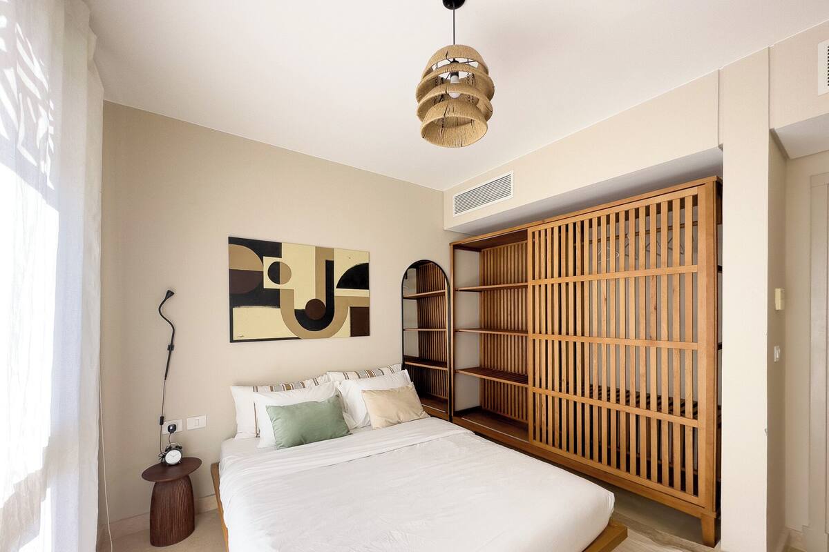 Een slaapkamer met een bed, een houten garderobe, een abstract schilderij aan de muur en een hanglamp.