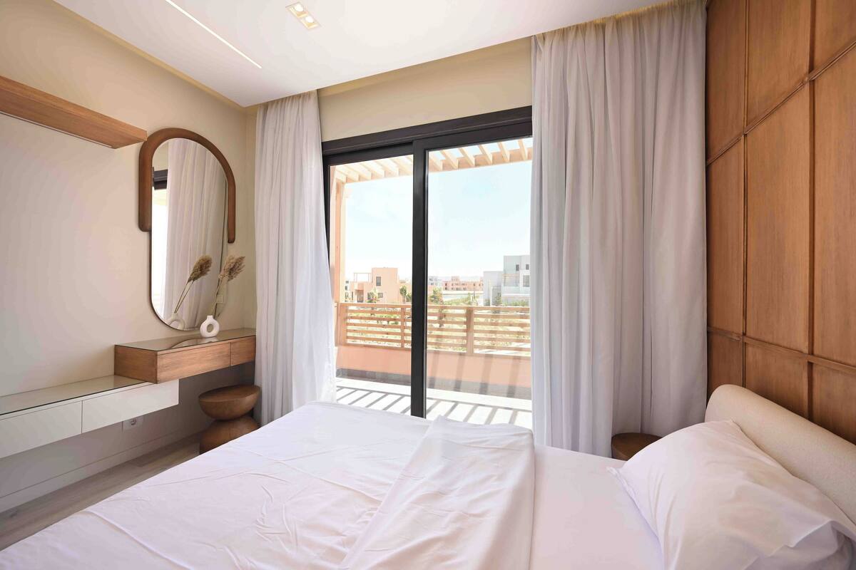 Een moderne hotelkamer met een groot bed, een houten hoofdbord, een groot raam met gordijnen, een houten wastafel met spiegel en uitzicht op de stad.
