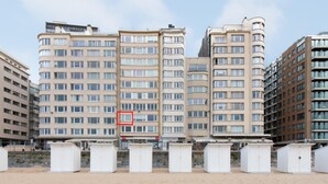 Una hilera de edificios residenciales altos con varios pisos y balcones, un terreno arenoso al frente y un cielo despejado.