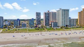 Ein Hotel direkt am Strand mit mehreren Hochhäusern, einem Sandstrand mit Sonnenschirmen und blauem, wolkenlosem Himmel.