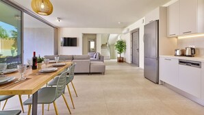 Eine moderne Küche mit integriertem Kühlschrank, ein Essbereich mit Holztisch und -stühlen sowie ein Wohnbereich mit Sofa und Fernseher.
