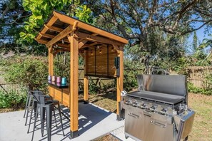 An outdoor barbecue area with a wooden pergola, a stainless steel grill, and a table with chairs.