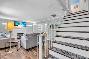 A modern living room with a fireplace, a staircase with decorative railings, and a large painting of a beach scene.