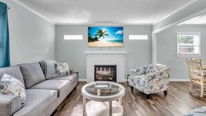 A living room with a gray sofa, a patterned armchair, a round coffee table, a fireplace, and a beach-themed painting above the fireplace. - Heated Salt Water Pool & Fire Pit Retreat in Sarasota (Sarasota)