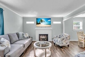 A living room with a gray sofa, a patterned armchair, a round coffee table, a fireplace, and a beach-themed painting above the fireplace.