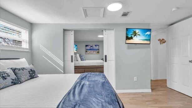 A bedroom with a bed, a nightstand, a window with blinds, a door leading to a bathroom, and a wall-mounted TV. - Heated Salt Water Pool & Fire Pit Retreat in Sarasota (Sarasota)