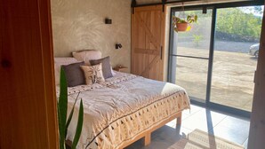 Un dormitorio con cama, almohadas, una planta colgante y vistas a través de un ventanal grande.