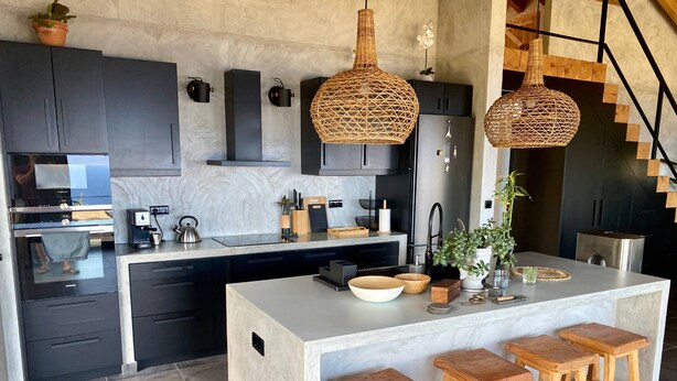Una cocina moderna con armarios oscuros, una isla grande con taburetes y lámparas colgantes de mimbre.