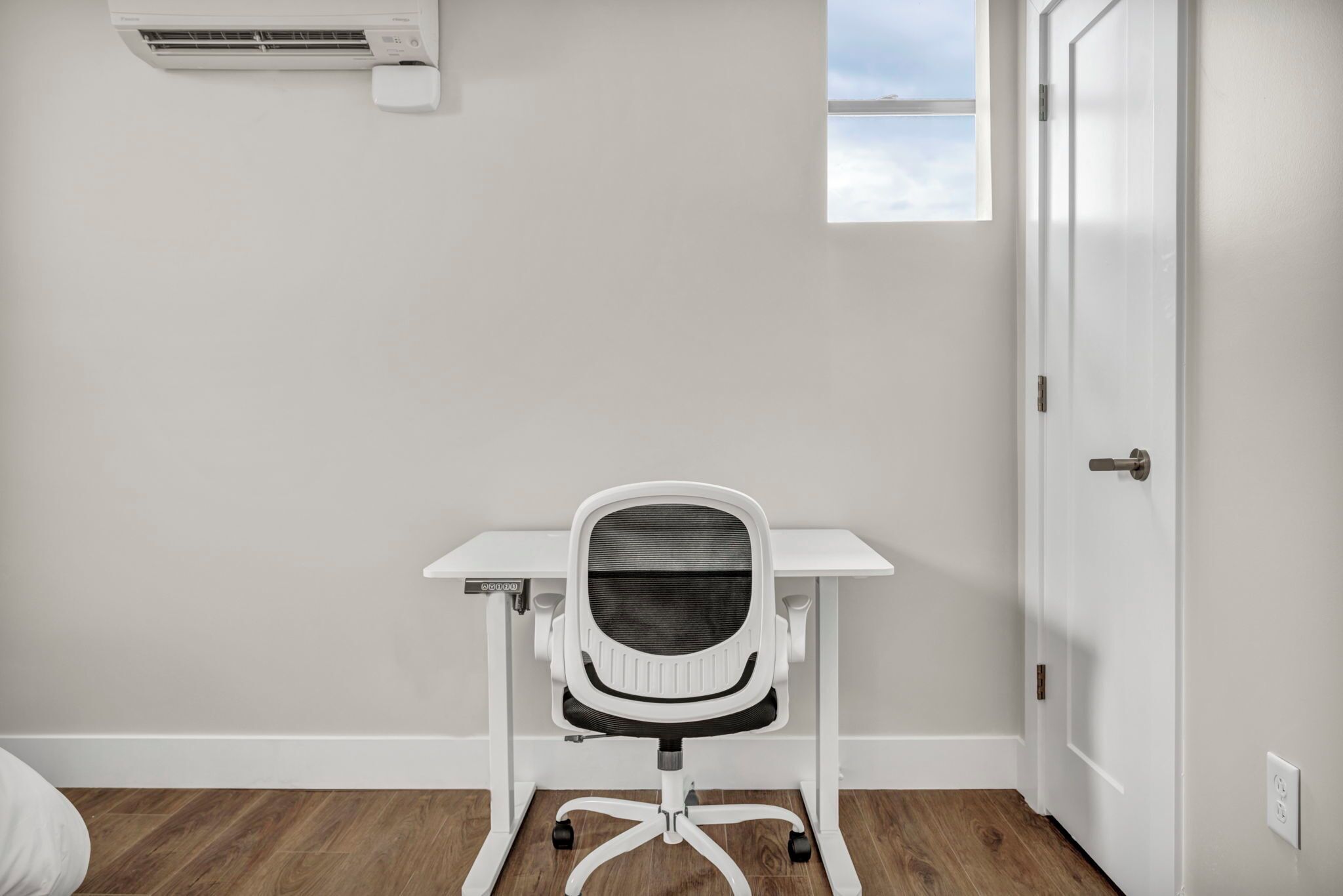 Une pièce avec un bureau blanc, une chaise de bureau blanche, une fenêtre donnant sur un paysage agréable et une porte.