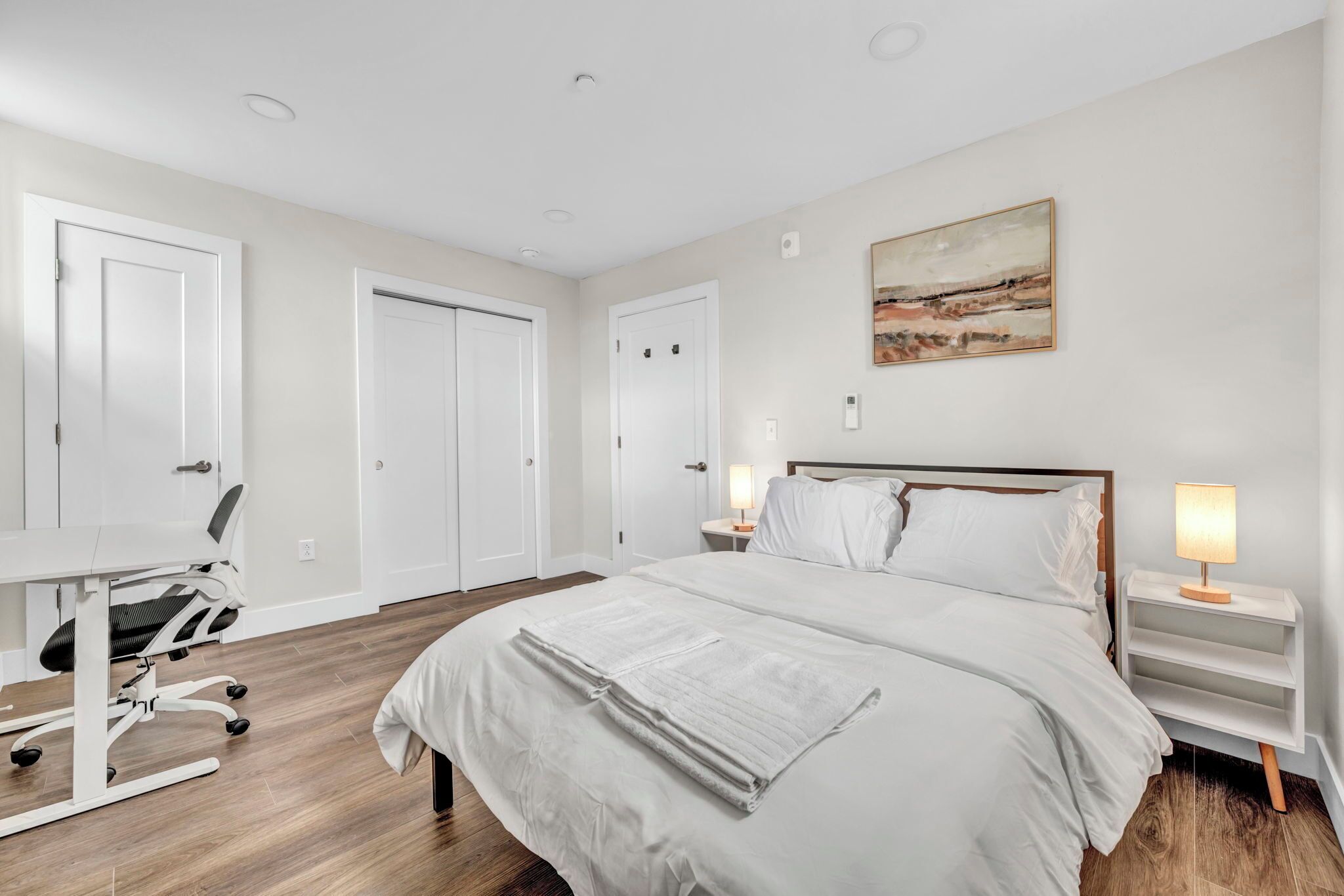 A hotel room with a large bed, a desk with a chair, two bedside tables with lamps, and a painting on the wall.