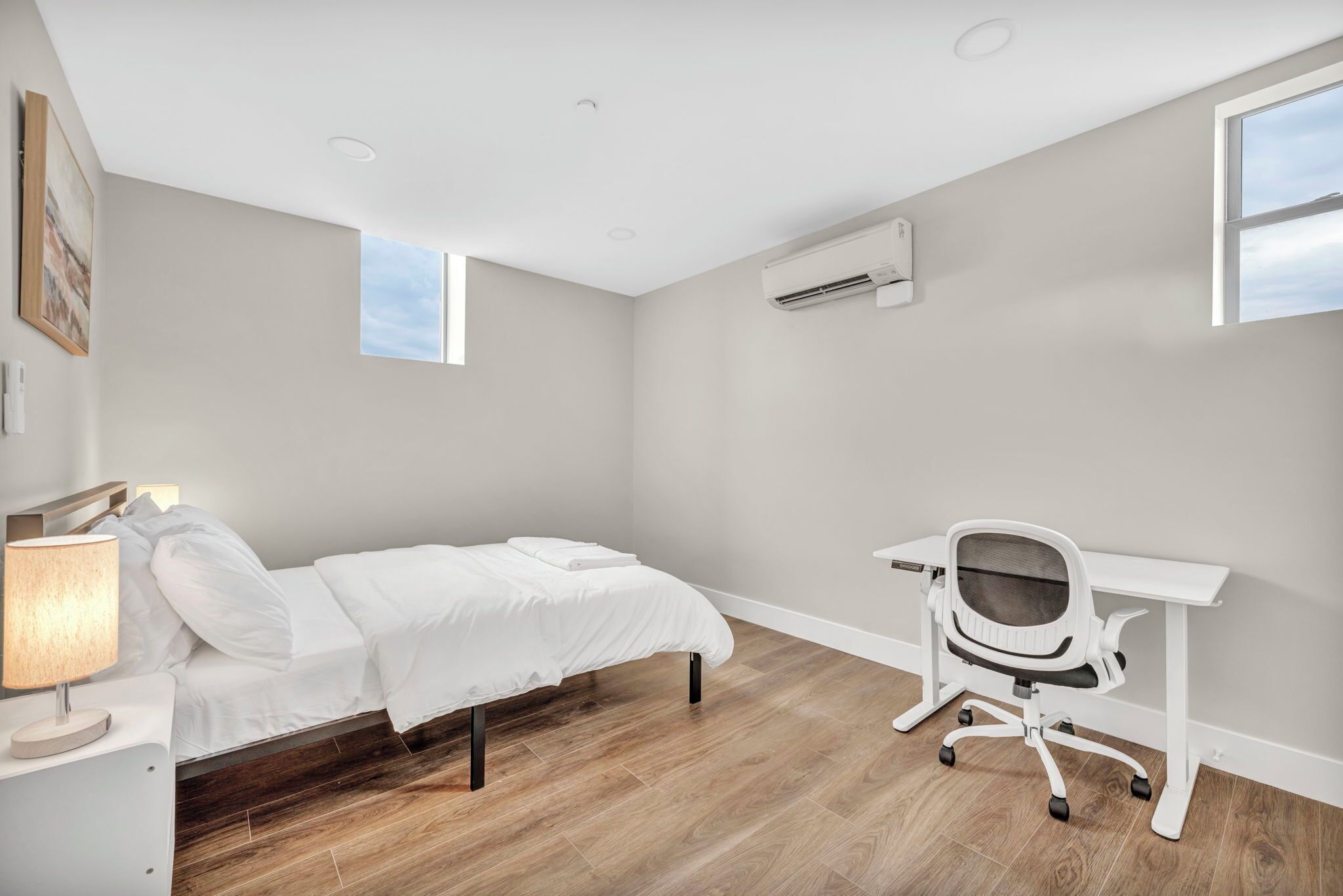 A bedroom with a bed, bedside table, lamp, desk, chair, and an air conditioning unit.