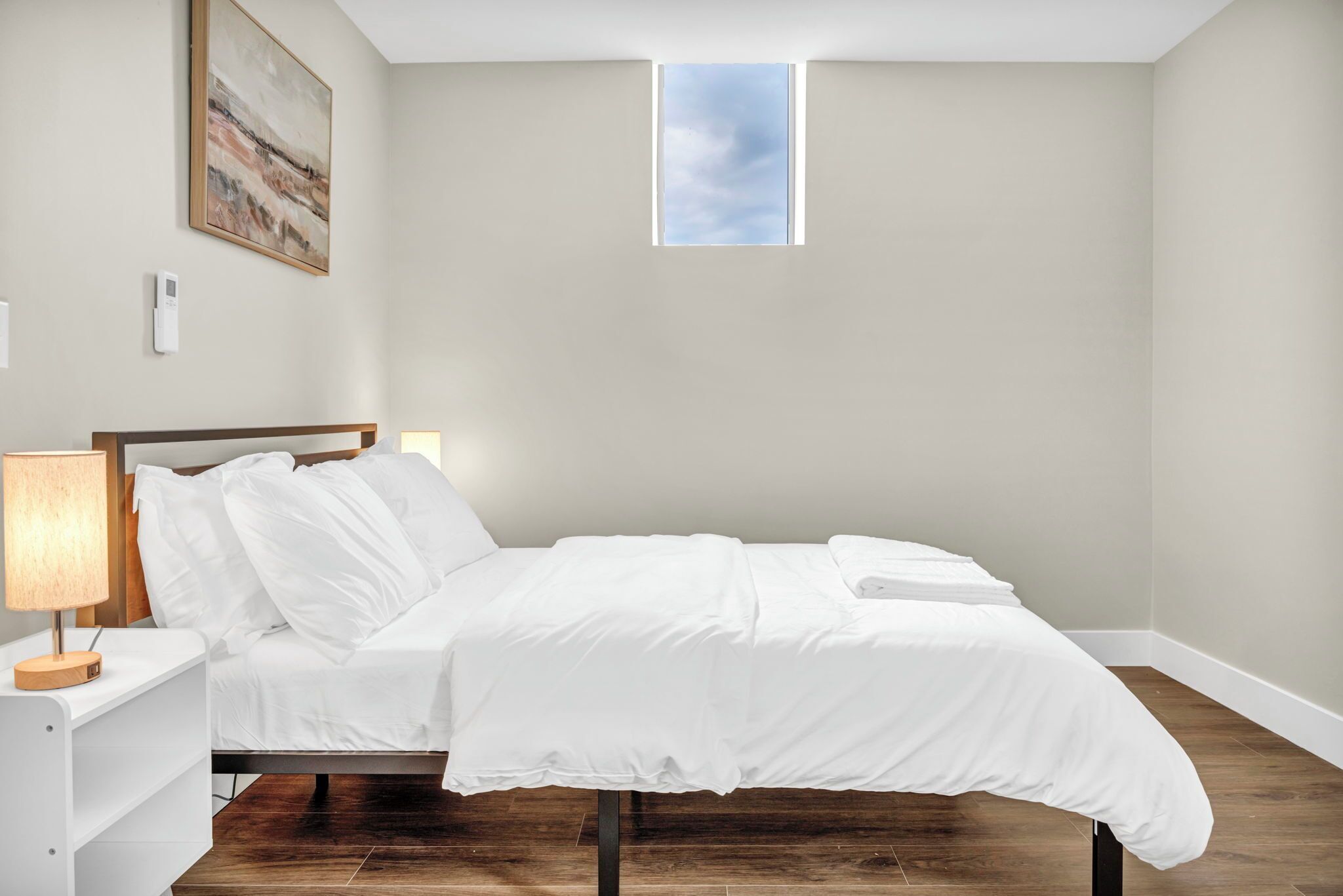 A neatly made bed with white bedding, a bedside table with a lamp, a small window, and a painting on the wall.