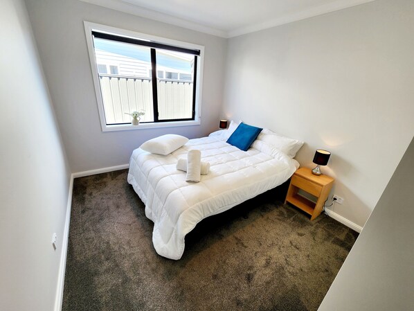A neatly made bed with white bedding and a blue pillow, a bedside lamp, and a window with blinds.