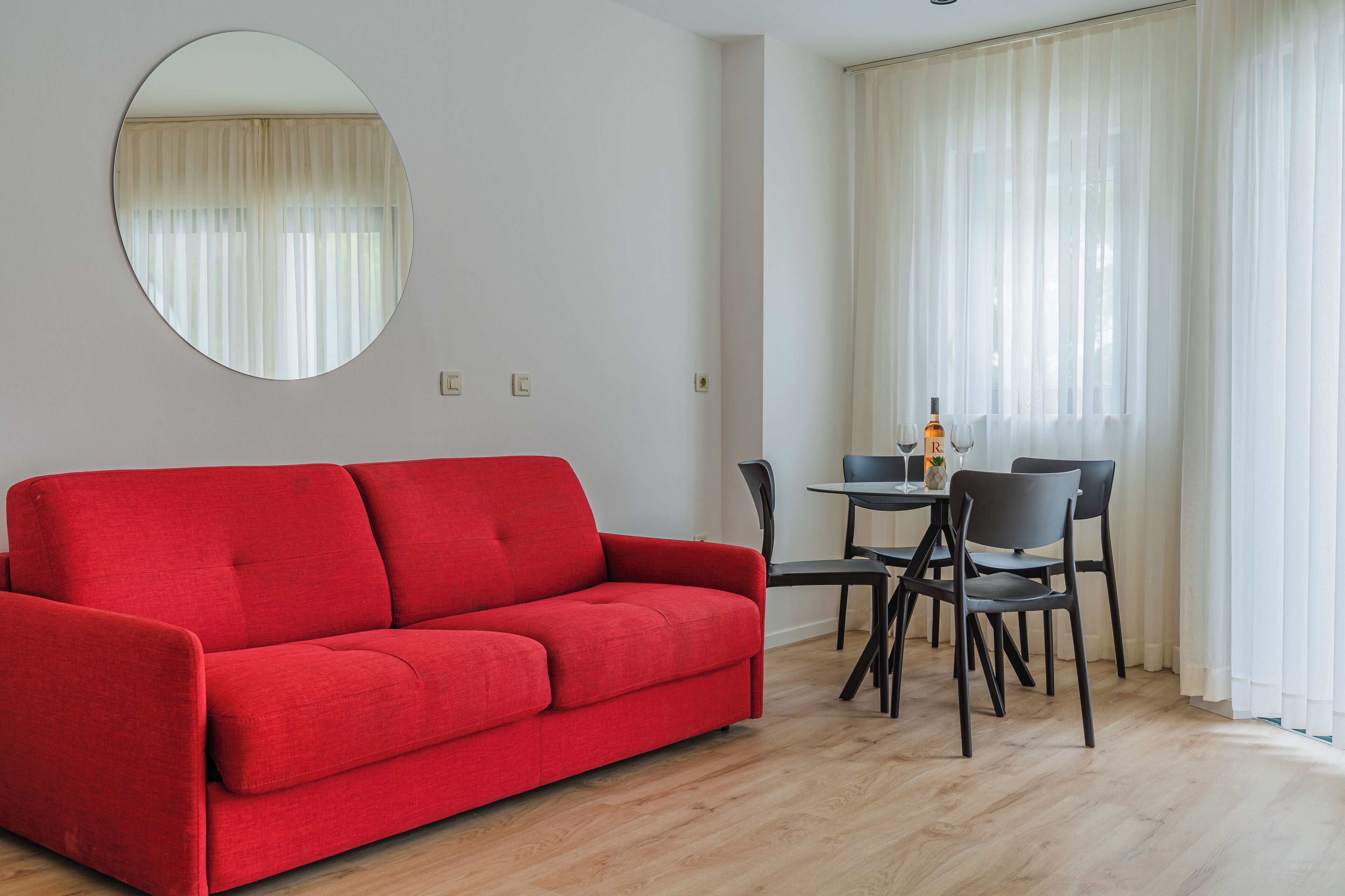 Ein rotes Sofa, ein runder Spiegel, ein Esstisch mit Stühlen und leichte Vorhänge.