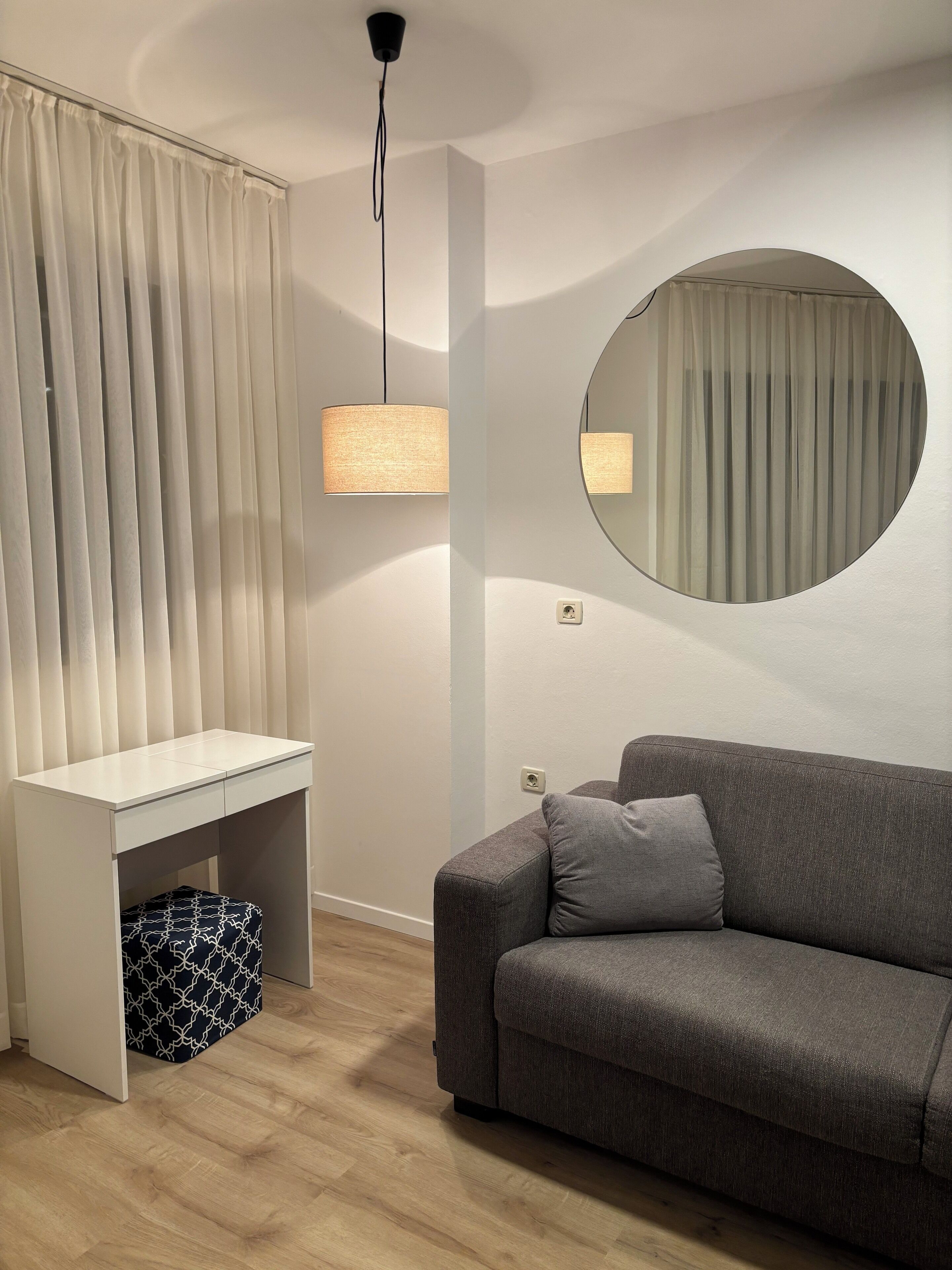 A modern room with a grey sofa, a white desk with a patterned ottoman, a round mirror, and a ceiling light.