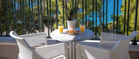 A balcony with white wicker chairs, a small round table with a potted plant, two glasses of orange juice, and a plate of pastries.