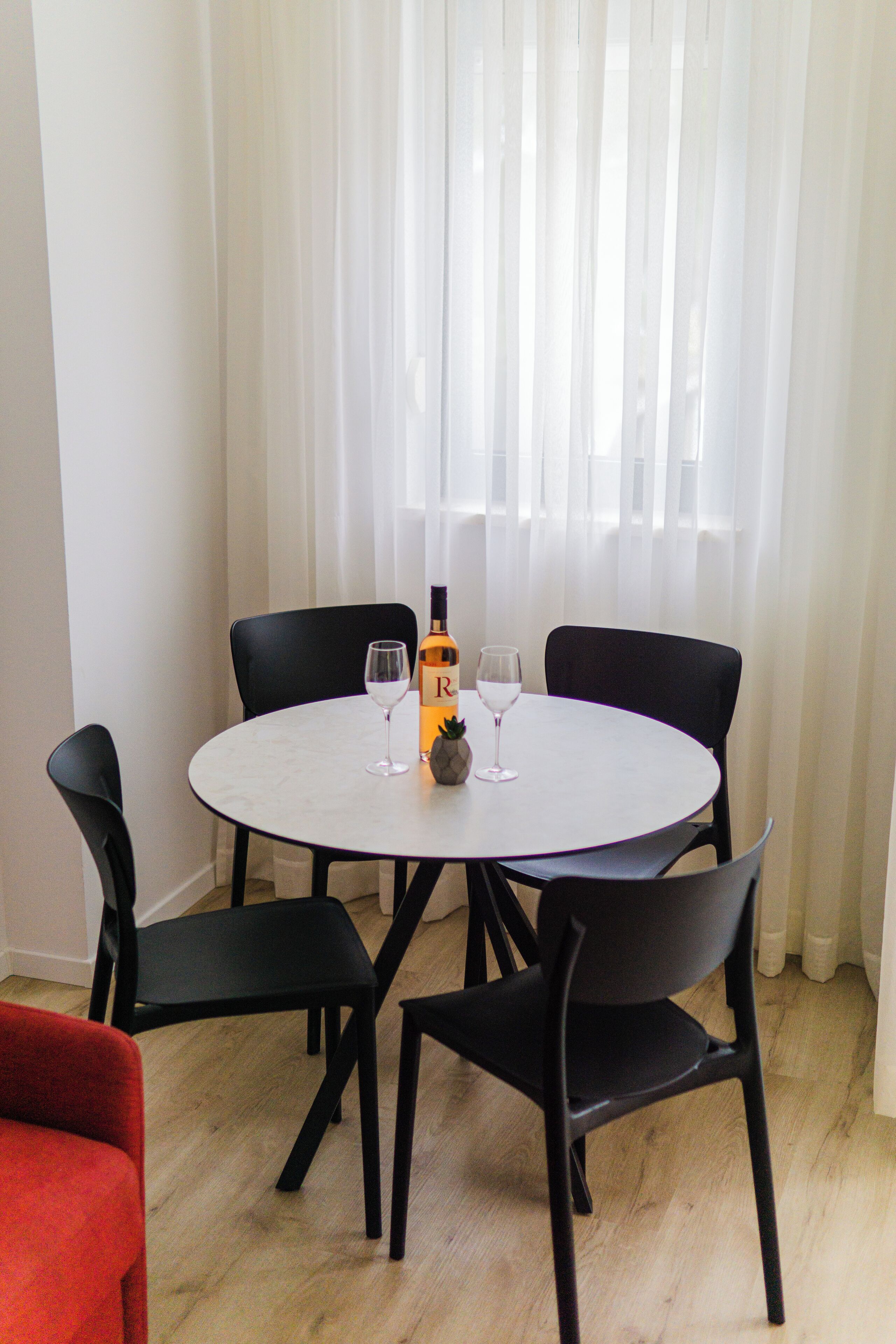 A round table with a bottle of wine and two glasses, surrounded by black chairs, in a room with light wooden flooring and white curtains.