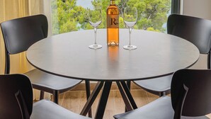 Ein runder Tisch mit vier Stühlen, einer Weinflasche und zwei Weingläsern. Der Tisch steht in der Nähe eines Fensters mit Blick auf Bäume und das Meer.