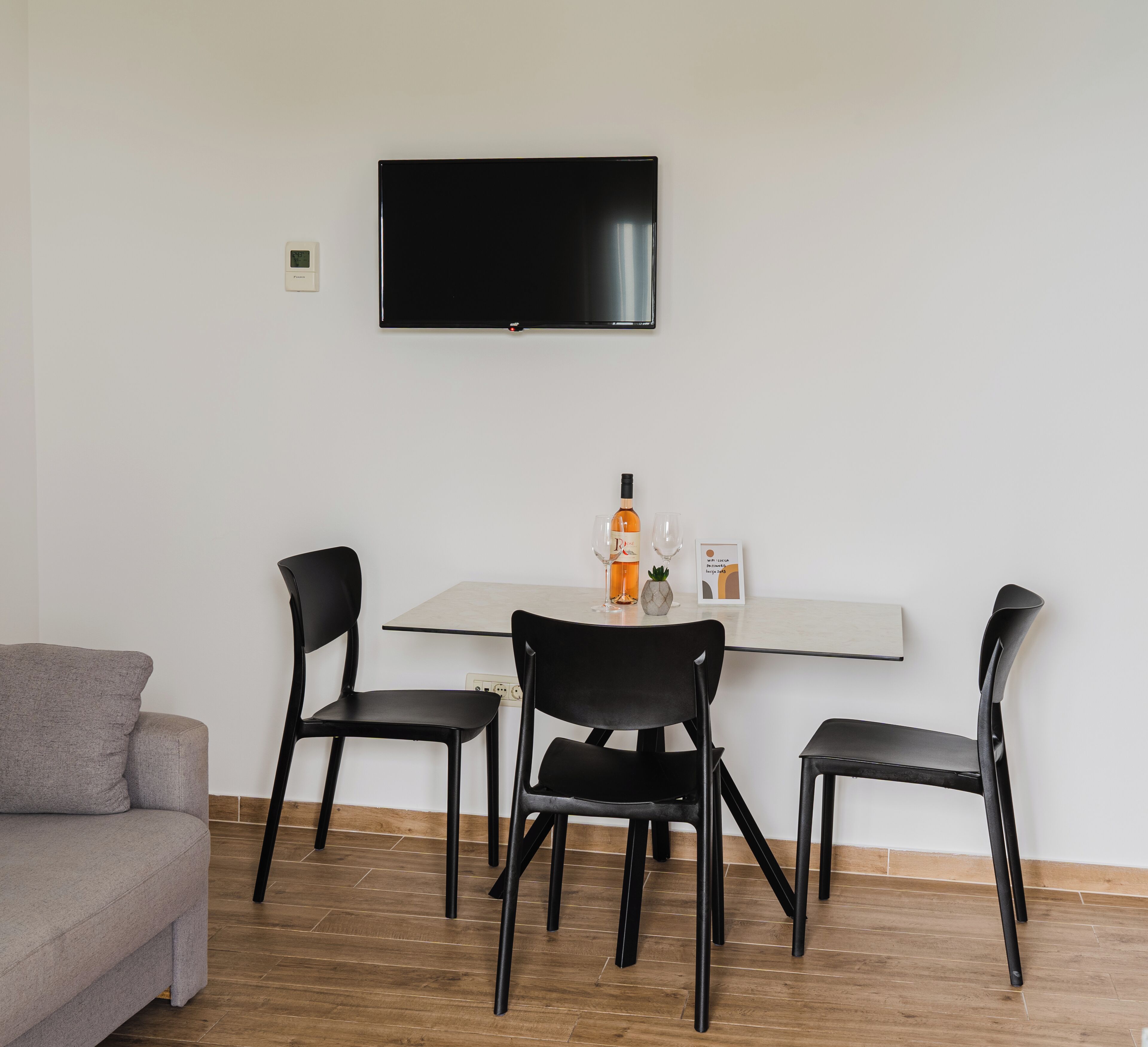 Un salon moderne avec un canapé gris, un plancher en bois, un téléviseur à écran plat fixé au mur et une table à manger avec des chaises noires.