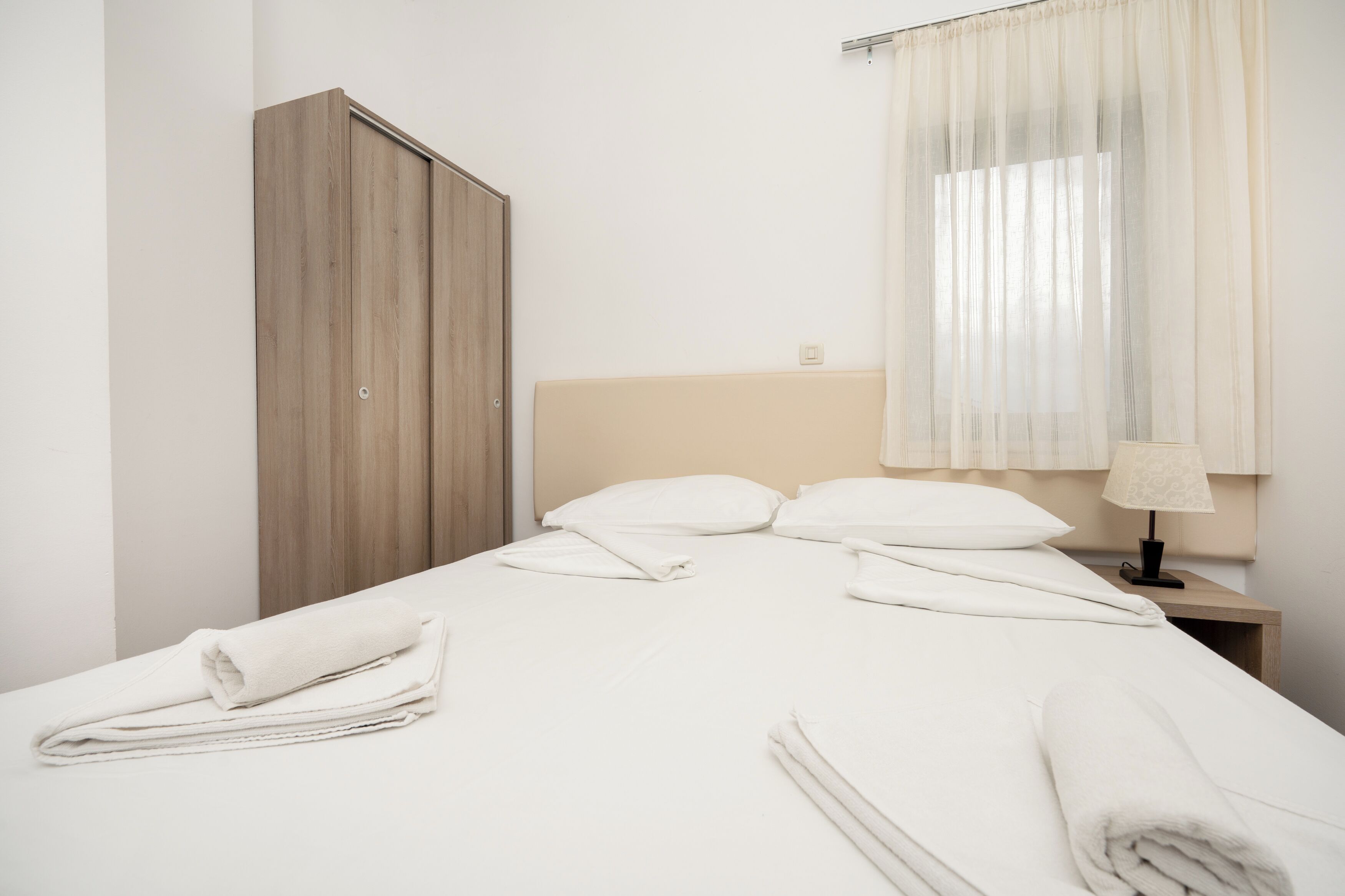 Ein ordentlich bezogenes Bett mit weißen Leinen und Handtüchern, ein Holzschrank, ein Nachttisch mit Lampe und ein Fenster mit durchsichtigen Vorhängen.