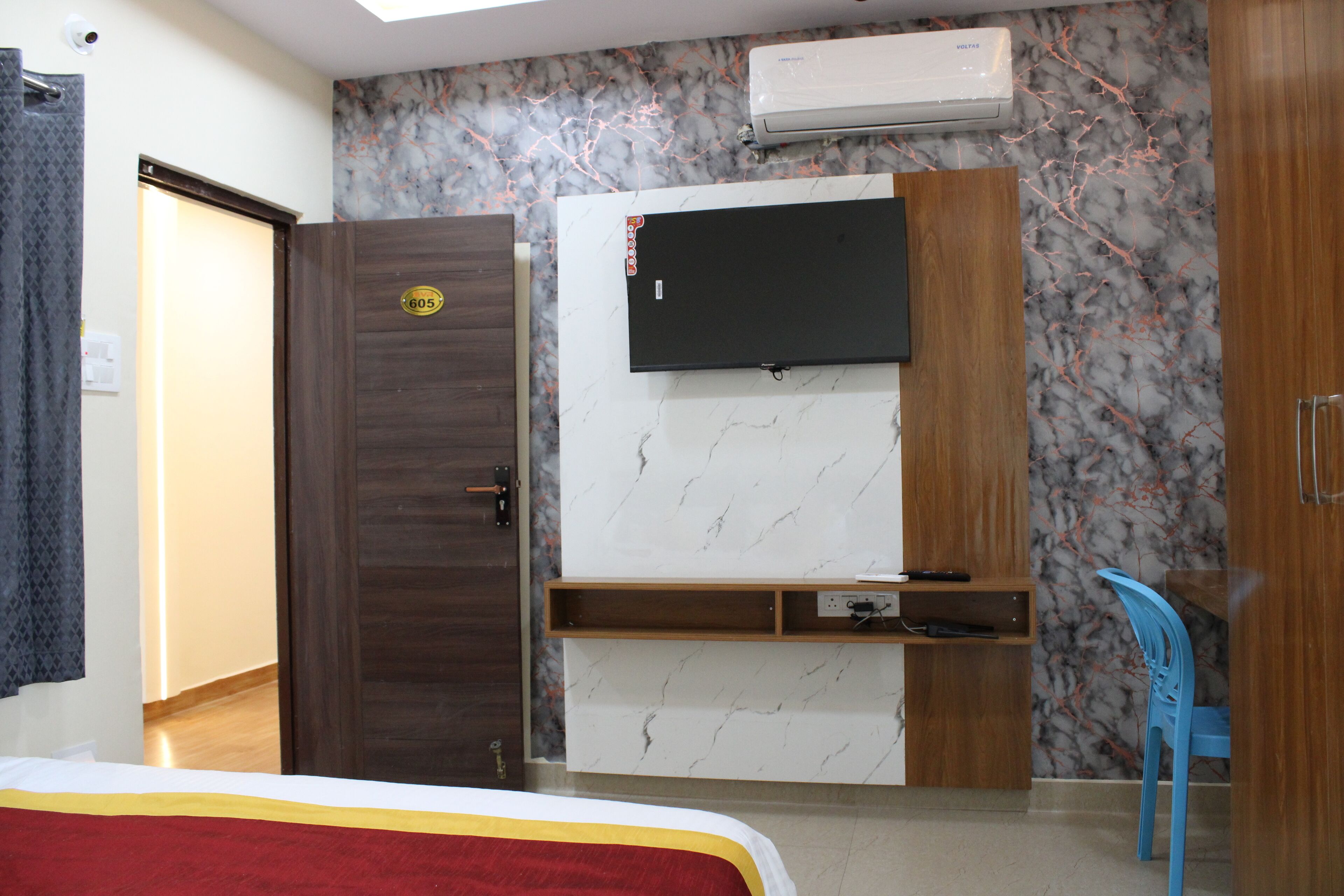 Une chambre d’hôtel avec un lit, un téléviseur fixé au mur en marbre, une armoire en bois et une chaise bleue.